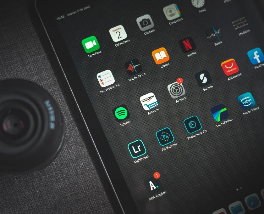 Uma tela de tablet exibindo uma variedade de ícones de aplicativos, incluindo FaceTime, Câmera, Netflix, Spotify, Amazon e Lightroom, entre outros. No canto esquerdo inferior, há uma parte visível de uma lente de câmera.